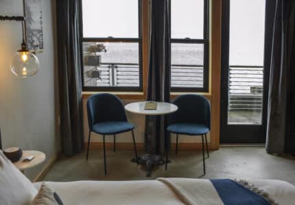 A cozy room features a bed in the foreground with white linens. Near the windows are two blue chairs and a small round table with a book. A glass pendant light hangs on the left. Outside, a foggy view and railing are visible through the windows.