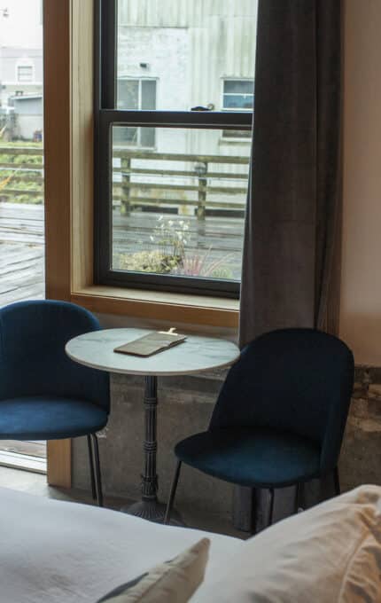 A cozy bedroom with a neatly made bed, two dark blue chairs, and a round table next to a window. A pendant light hangs overhead. The window overlooks a wooden deck with plants, and a glass door provides access to the outside.