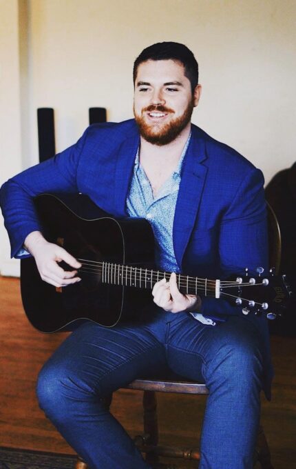 A man with a beard is seated on a wooden chair, playing a black acoustic guitar. He is wearing a blue suit and a light blue patterned shirt. The background is a simple room with wooden flooring and a beige wall.