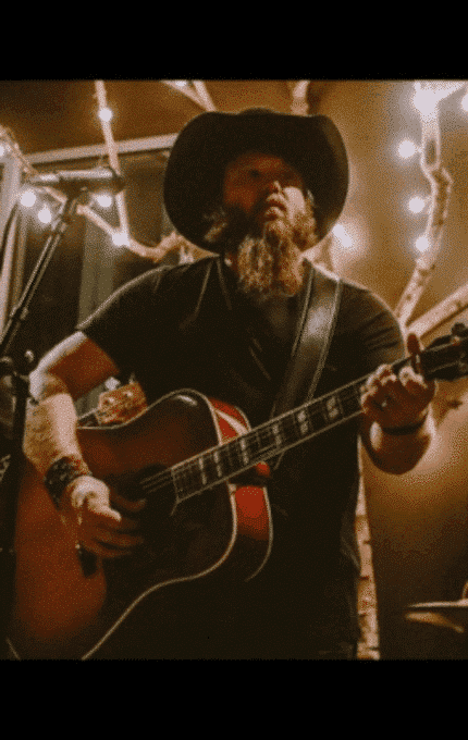 A man with a beard and cowboy hat plays an acoustic guitar and sings into a microphone. He is dressed in dark clothing and is surrounded by string lights in a dimly lit room.