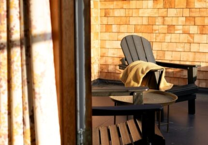 Two wooden Adirondack chairs and a small round table are on a porch with wood shingle walls. A yellow blanket is draped over one chair. Sunlight streams in from the left, partially illuminating the scene.
