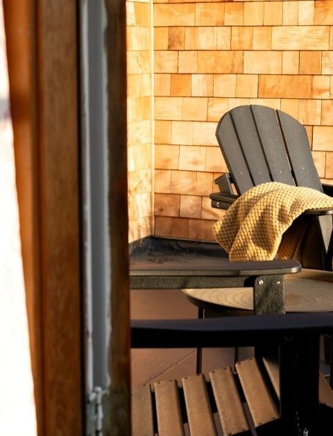 Two wooden Adirondack chairs and a small round table are on a porch with wood shingle walls. A yellow blanket is draped over one chair. Sunlight streams in from the left, partially illuminating the scene.