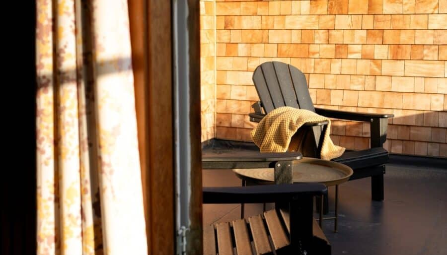 Two wooden Adirondack chairs and a small round table are on a porch with wood shingle walls. A yellow blanket is draped over one chair. Sunlight streams in from the left, partially illuminating the scene.
