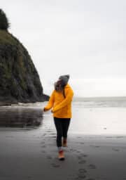 A person wearing a yellow jacket and gray beanie is walking on a beach, leaving footprints in the sand. The ocean and a rocky cliff are in the background under an overcast sky.