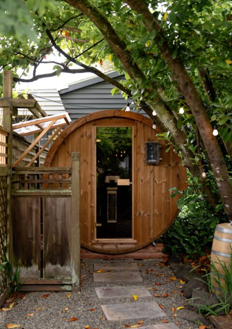 A barrel-shaped wooden sauna sits in a garden, accessed by a stone path. Trees and plants surround the sauna, and a wooden fence is on the left side. There is a glass door with a wooden handle in the center.