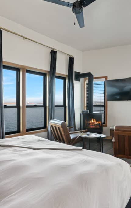 A bedroom with a large bed and dark curtains overlooks an ocean view through wide windows. The room features a wall-mounted TV, armchairs by a small table, a fireplace, and a ceiling fan. Dark wood flooring and a simple framed picture are also visible.