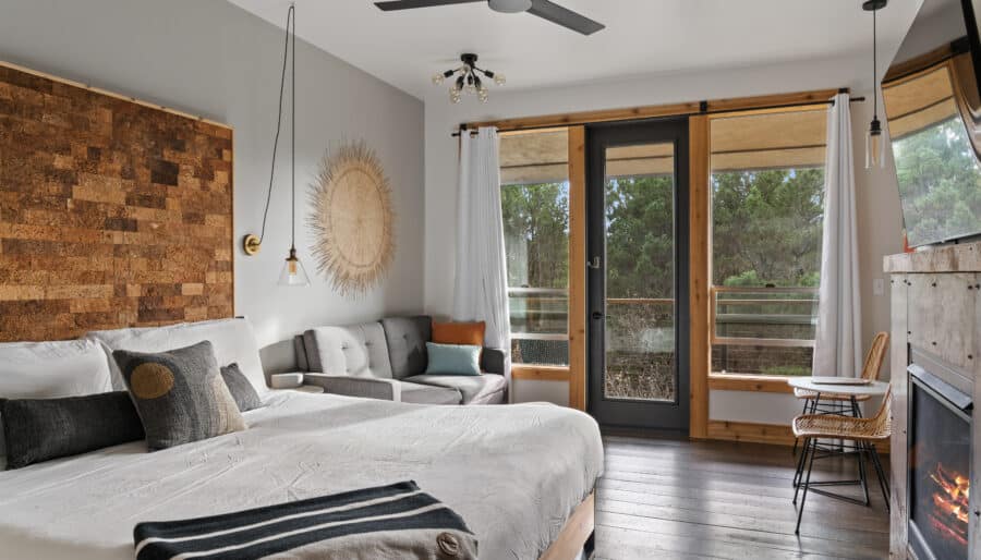 A cozy bedroom with a large bed featuring white and gray bedding. There's a cork headboard, a gray sofa, and a small dining table by a window. A fireplace is lit on the right wall. The room opens to a balcony with a view of trees outside.