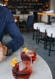 Two glasses containing a dark red cocktail with ice and a lemon twist garnish sit on a round marble table. A person's arm in a blue long-sleeve shirt and watch is visible on the left. The background shows a bar area with chairs and stools.