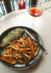 A plate with steak topped with greens, accompanied by fries, is on a table. A knife rests on the edge of the plate. A cocktail glass filled with a reddish drink is placed on the right side of the plate. A stained glass window is in the background.