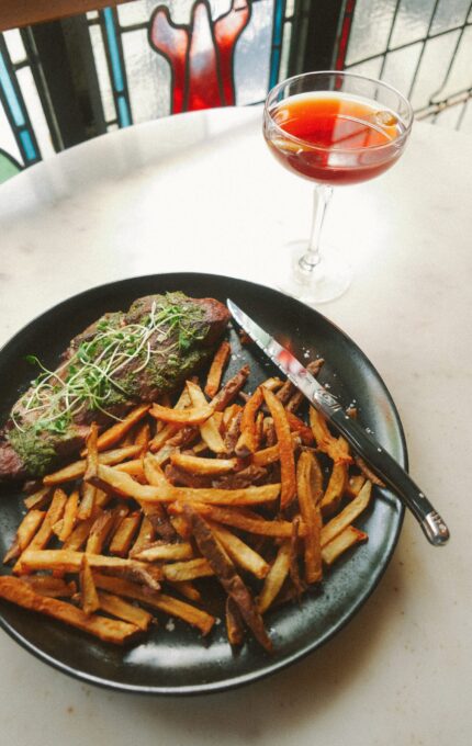A plate with steak topped with greens, accompanied by fries, is on a table. A knife rests on the edge of the plate. A cocktail glass filled with a reddish drink is placed on the right side of the plate. A stained glass window is in the background.
