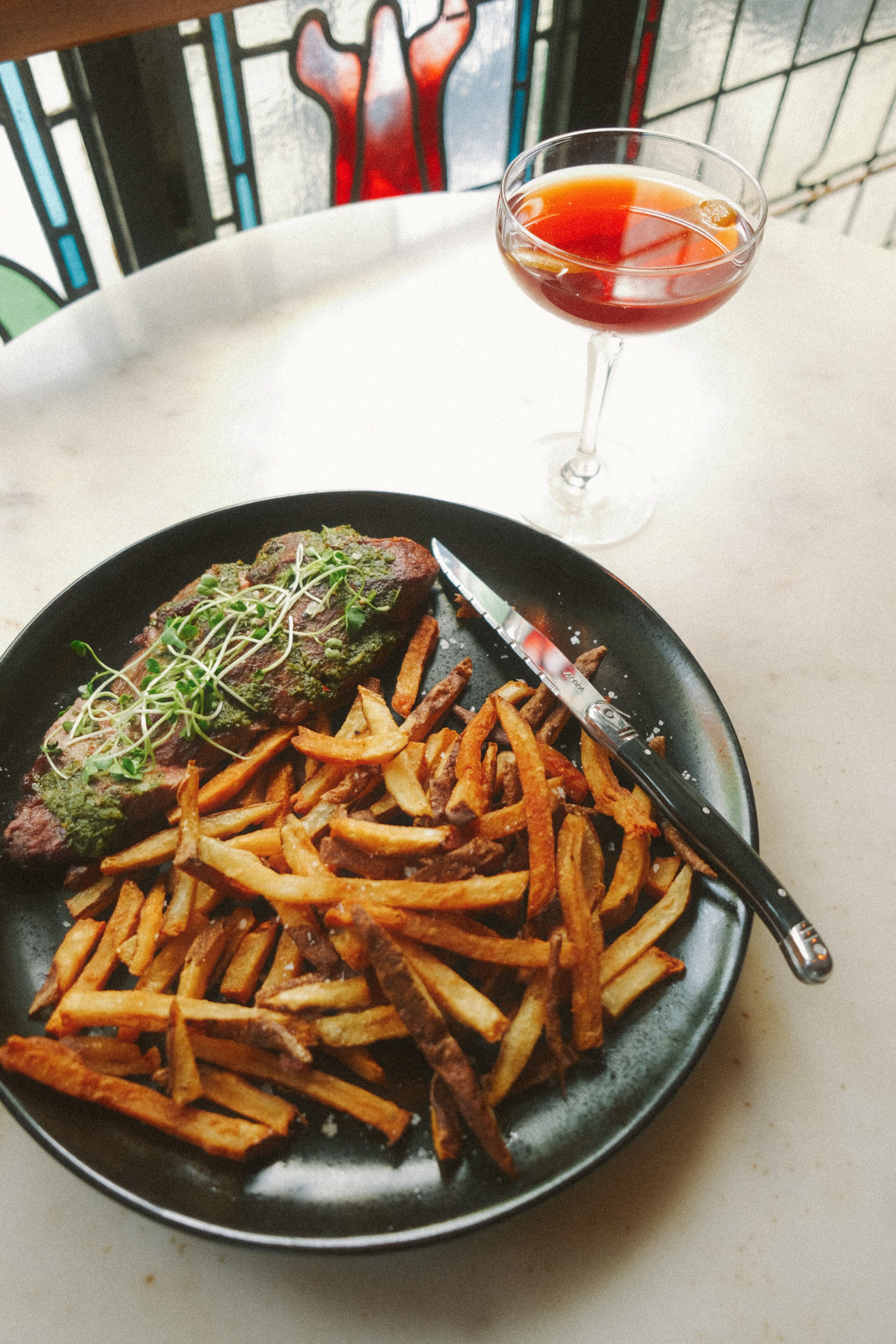 A plate with steak topped with greens, accompanied by fries, is on a table. A knife rests on the edge of the plate. A cocktail glass filled with a reddish drink is placed on the right side of the plate. A stained glass window is in the background.