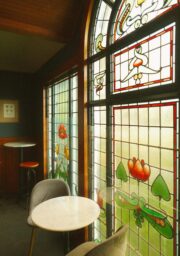 A cozy seating area with small white tables and gray chairs is illuminated by natural light from large stained glass windows featuring abstract floral designs in red, green, and orange hues. A framed artwork hangs on the wall.