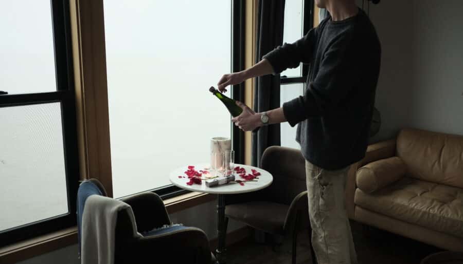 A person stands by a round table next to large windows, pouring a bottle of champagne into glasses. The table is scattered with rose petals and two empty glasses. There is a beige couch and chairs in the room.
