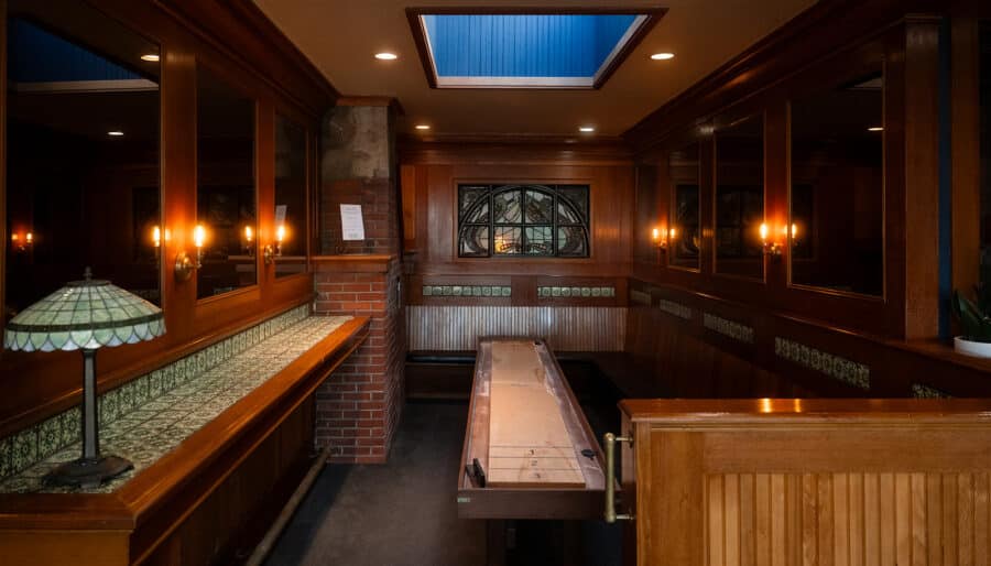 A narrow, wood-paneled room with a shuffleboard table in the center, stained glass window, cushioned bench seating, and warm lighting from wall sconces and a decorative table lamp.