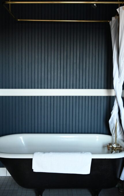 A bathroom with dark blue vertical paneling, a freestanding black clawfoot bathtub, white curtain partially pulled to one side, white towels on racks, and toiletries on a small round table beside the tub.