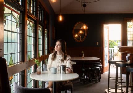 A woman sits alone at a table in a dimly lit café with stained glass windows, holding a mug and looking outside. There is a coffee pot on the table and an open door in the background letting in natural light.