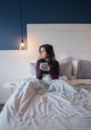 A person sits on a bed holding a mug, looking to the side. The room has a dark blue wall, a white wooden headboard, and striped bedding. A hanging light bulb is turned on above a small round bedside table.