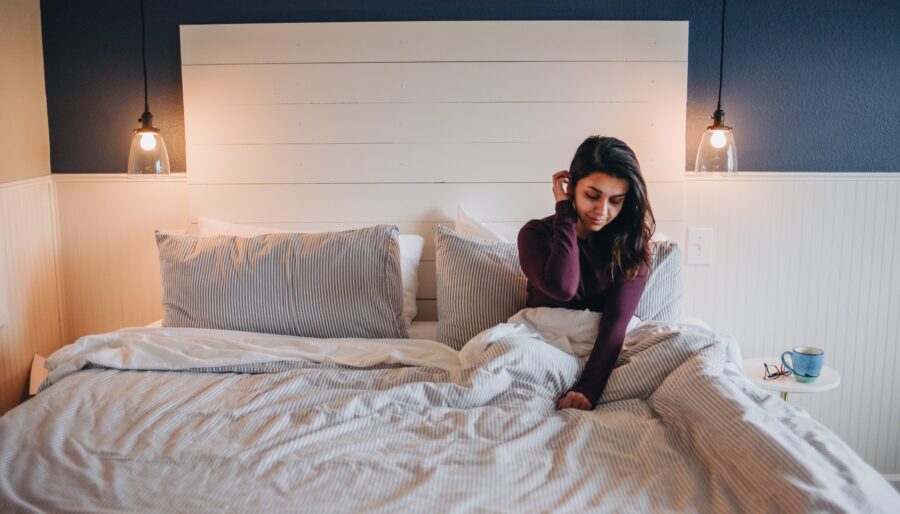 A person sits on a bed with gray and white striped bedding, under two hanging light bulbs, against a dark blue wall. A pair of glasses and a mug sit on a bedside table. The person is looking down, touching their hair.
