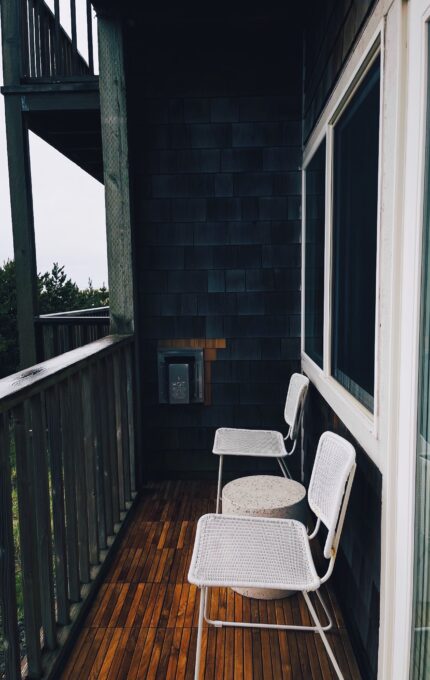 A wooden deck with two white metal chairs and a small round table is seen attached to a dark shingled building. A sliding glass door is on the right, and there is greenery visible beyond the deck's wooden railing.