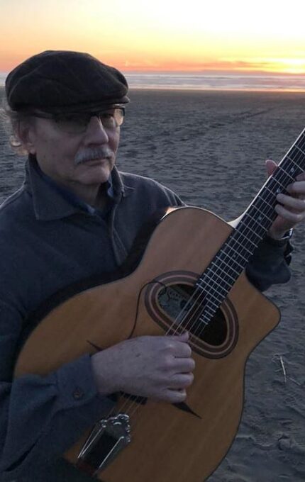 An older man wearing a cap and glasses is playing a guitar on a sandy beach during sunset. In the background, the ocean is visible, and a few people are walking along the shore.