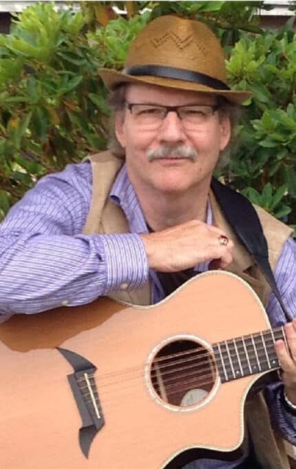 A man wearing a straw hat, glasses, and a striped shirt sits outdoors, holding an acoustic guitar. There are green bushes in the background.