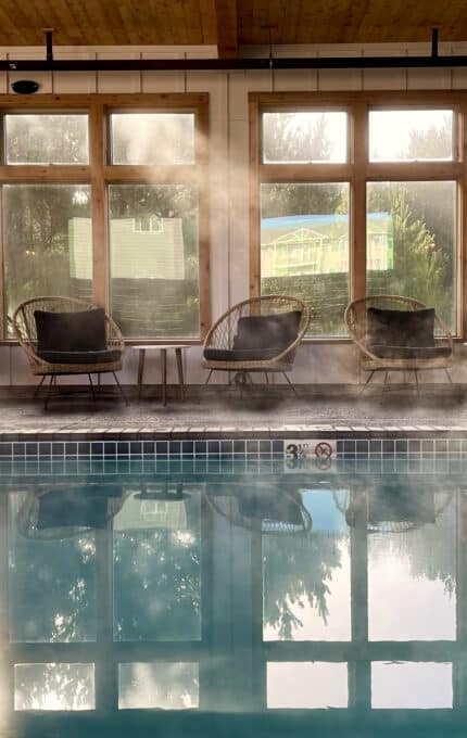 Indoor pool area with steam rising from the water. The room has large windows and several chairs arranged along the wall. The ceiling is wooden, and the pool area is well-lit with natural light. The depth markers indicate three to four feet.