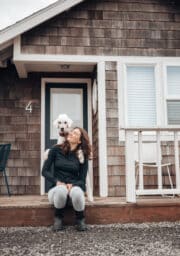 A woman sits on the wooden steps of a house with shingle siding, smiling while a large white dog playfully places its front paws on her shoulders. House number 4 is visible next to the door. A chair is positioned near the entrance.