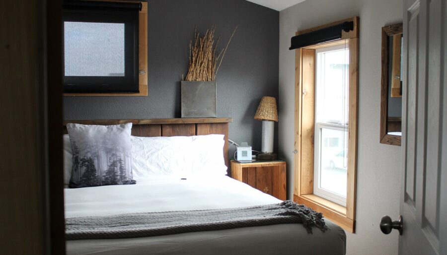 A small bedroom featuring a bed with white linens and decorative pillows, a wooden headboard, and a side table with a lamp. The room has gray and white walls, a window with wooden trim, and simple decor.