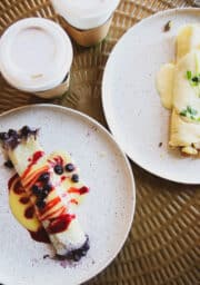 Two plates with crepes sit on a textured surface. One crepe is topped with blueberries and red syrup, while the other has a creamy sauce and garnish. Two coffee cups with lids are in the background.