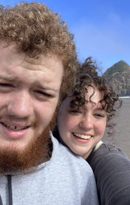 A man with curly hair and a beard wearing a grey hoodie and a woman with curly hair in a grey sweater are standing on the beach. They are smiling with a large rock formation and blue sky in the background.