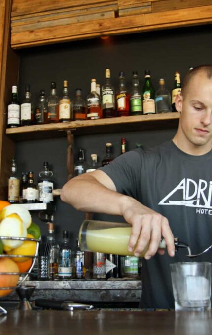 A bartender wearing a gray "Adrift Hotel + Spa" t-shirt is seen behind a bar counter pouring a drink from a small pitcher into a glass filled with ice. The counter has a basket of assorted fruits, and shelves of various bottled alcohol are visible in the background.