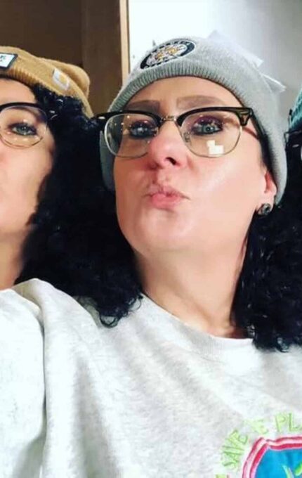 Three people with curly hair are posing for a selfie indoors, all wearing glasses, beanies, and casual clothing. They are making playful, pouty facial expressions and appear to be enjoying themselves.