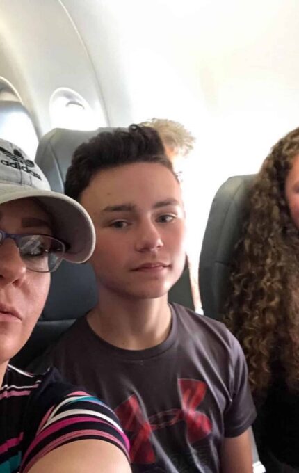 A woman, a boy, and a girl with curly hair are seated in an airplane. The woman is wearing glasses and a hat, the boy is wearing a dark shirt, and the girl is wearing a light-colored top. Empty seats are visible in the background.