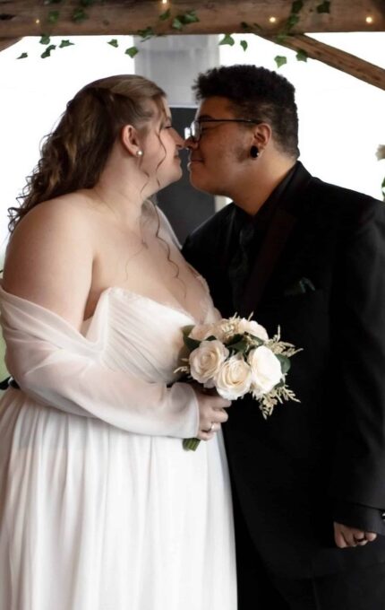 Two people dressed in formal attire are close together under a wooden arch draped with greenery and lights. One person is wearing a white dress and holding a bouquet of roses, while the other is wearing a black suit. They are about to kiss.