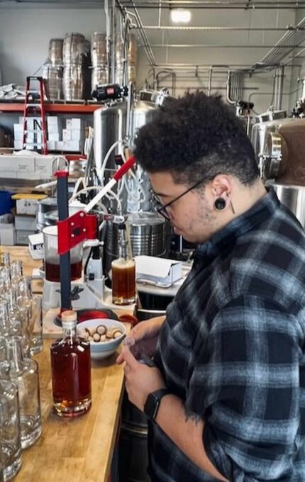 A person in a plaid shirt and glasses is working on a wooden table in a brewery. They are capping bottles with a red-handled bottle capper. Several bottles, caps, and a filled glass are on the table. Brewery equipment is visible in the background.