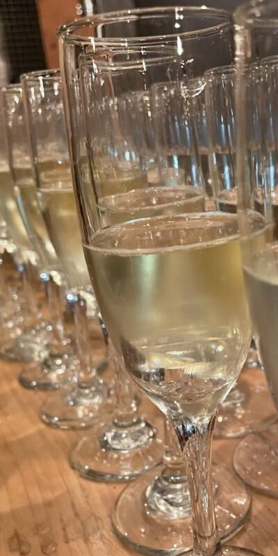 A row of filled champagne glasses is arranged on a wooden table. In the background, there are two bottles and a cork. The scene suggests a celebration or gathering.