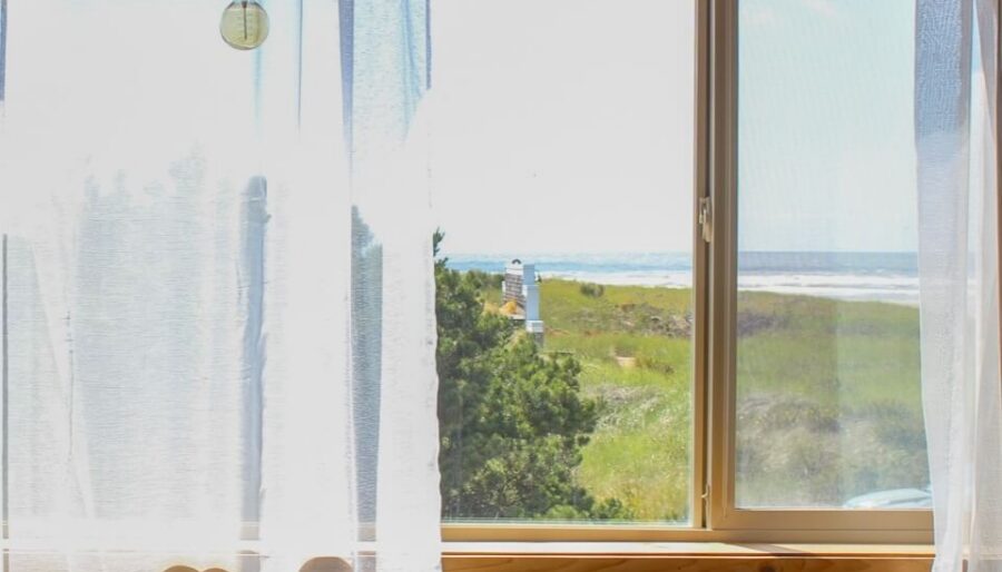 Two gray armchairs and a small round table are positioned in front of a window with sheer white curtains. Outside, green grass and a glimpse of water and blue sky are visible. A single light bulb hangs from the ceiling.