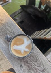 A ceramic cup of coffee on a wooden deck features latte art depicting a bird in flight. The cup is positioned near the edge of the deck, overlooking water below.