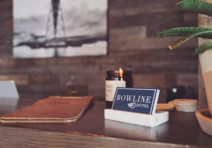 A reception desk with a "Bowline Hotel" sign. The background features a black and white framed photo, and there are various items on the desk, including a lit candle, a leather-bound book, and a potted plant.