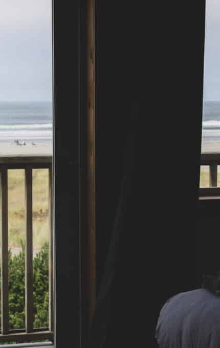 View from a dimly lit room through open sliding doors with curtains to a sandy beach and ocean. People are visible in the distance on the beach. A kite is flying above the shoreline. A bed partially visible in the foreground.