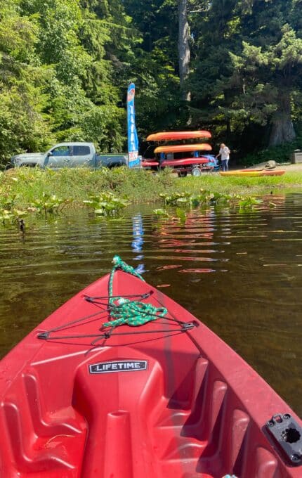 A red kayak floats near the edge of a calm body of water. In the background, vehicles, a rental sign, stacked kayaks, and two people are visible near trees and greenery.