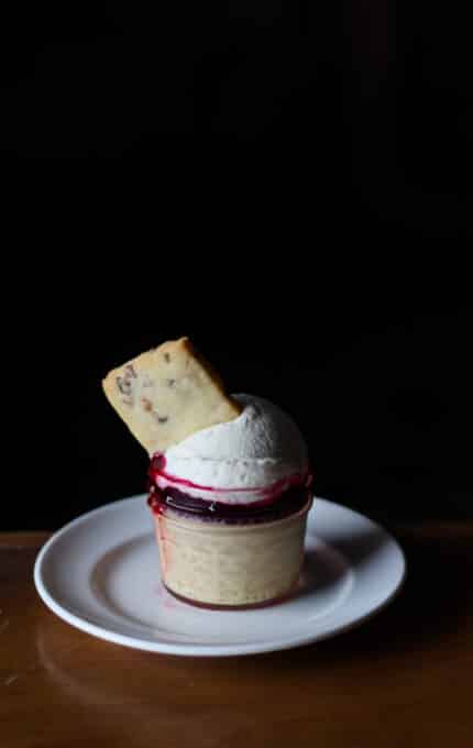 A dessert is presented on a white plate. It features a cream layer topped with a dollop of whipped cream and a red syrup, with a rectangular cookie inserted into the cream. The background is dark, highlighting the dessert.