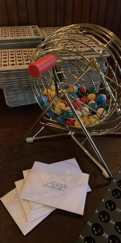 A bingo cage with colorful balls sits on a table. Behind it are stacks of bingo cards. To the side, a black tray with numbered slots and white envelopes labeled "Adrift" are visible.