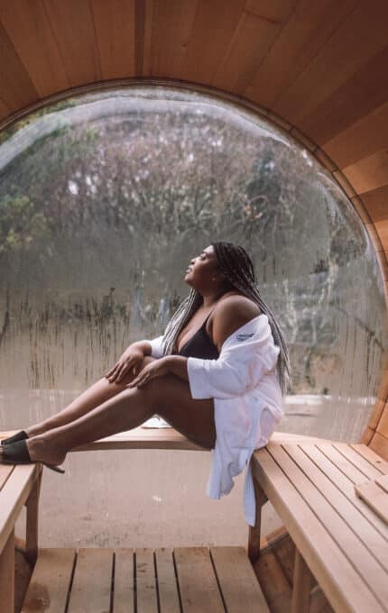 A woman with long braided hair sits on a wooden bench in a circular wooden sauna. She is wearing a white robe and looking up, with a misty window behind her, showing a blurred forest view.