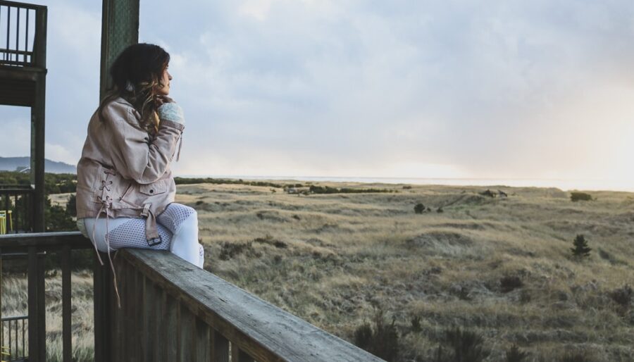 A person sits on a wooden railing, overlooking a vast field under a cloudy sky. They are wearing a light jacket and white pants, gazing thoughtfully into the distance. The scene appears peaceful and serene.