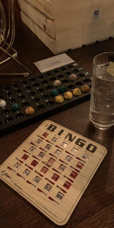A wooden table with a bingo card, a glass of clear drink with ice and a lime wedge, and a bingo cage with numbered balls and a tray holding selected balls.