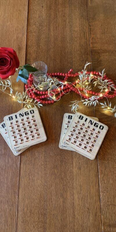 A wooden table displays bingo cards, a red rose in a small vase, red beaded necklaces, gold garland, and a string of white lights. The items are arranged decoratively across the surface.