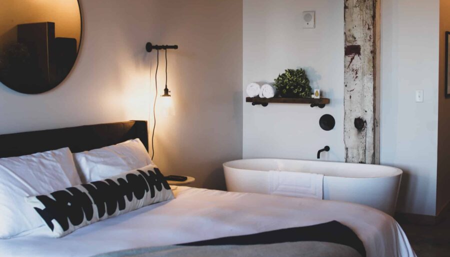 A modern bedroom features a round mirror, a bed with white and black pillows, and a side table with a lamp. There is a freestanding bathtub with folded towels and plants on a wooden shelf nearby. The room has neutral tones and soft lighting.