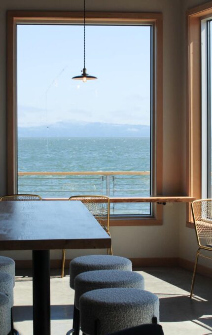 A cafe interior with large windows framing a view of the ocean. There are pendant lights hanging from the ceiling, a long wooden table with round stools in the center, and metal chairs with woven backs near the windows.