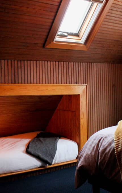 A cozy attic bedroom with wooden paneling features a single bed built into a nook with a reading light, a pillow, and a dark blanket, beside a larger bed with a yellow throw, under a skylight window.